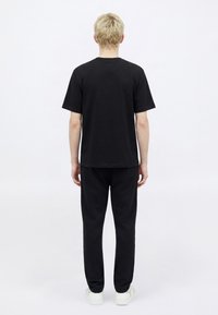 Fekete pamut rövid ujjú póló kerek nyakkivágással, hozzáillő fekete, szűkített fazonú nadrággal párosítva. A modellen fehér tornacipő látható.