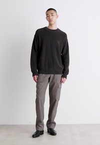 Maglione nero a coste con scollo rotondo e tasca laterale abbinato a pantaloni cargo grigi larghi. Il modello indossa scarpe basse nere, in piedi su uno sfondo chiaro.