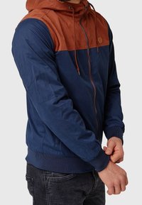 Homme portant une veste à capuche bleu marine et couleur rouille, ajustant le poignet de la veste avec ses mains.