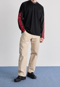 Person wearing black Adidas sweatshirt with red stripes, beige pants, and black sneakers standing on blue carpet against white wall.