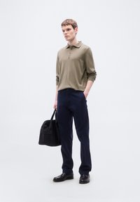 Πράσινο χακί μακρυμάνικο πόλο πουκάμισο, μπλε σκούρο παντελόνι, μαύρα παπούτσια, κρατώντας μια μαύρη τσάντα. Απλός, εφαρμοστός σχεδιασμός με γραμμή κουμπιών.