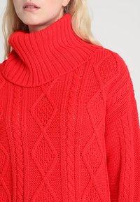 Červený pletený rolák se strukturovanými vzory ve tvaru diamantů a copánků. Má široký žebrovaný límec a volný střih.