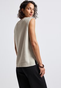 Kvinna med lockigt hår som bär en beige ärmlös stickad topp och svarta byxor, ser över sin axel mot en enfärgad bakgrund.