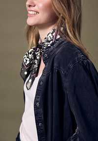 Camicia in denim nera con bottoni argentati, indossata sopra una maglietta bianca, con una sciarpa a motivi bianchi e neri annodata al collo.
