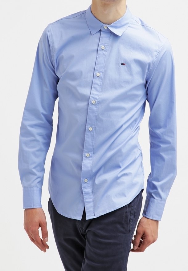 Homme portant une chemise bleu clair à manches longues avec un petit logo sur la poitrine et un pantalon sombre sur un fond uni.