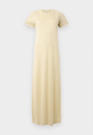 Femme debout, main sur la hanche, portant une robe maxi jaune pâle à manches courtes et des sandales à talons noires sur un fond uni.