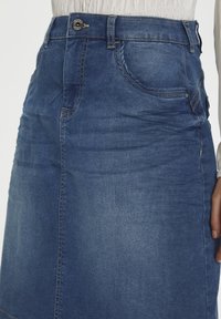 Jeanskjol i mörkblått, med en figurnära midja, framfickor och kontrasterande sömmar. Materialet har en slät, lätt texturerad yta.