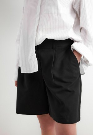 Personne portant un short noir sur mesure et une chemise blanche à manches longues retroussées, main dans la poche, sur un fond uni.