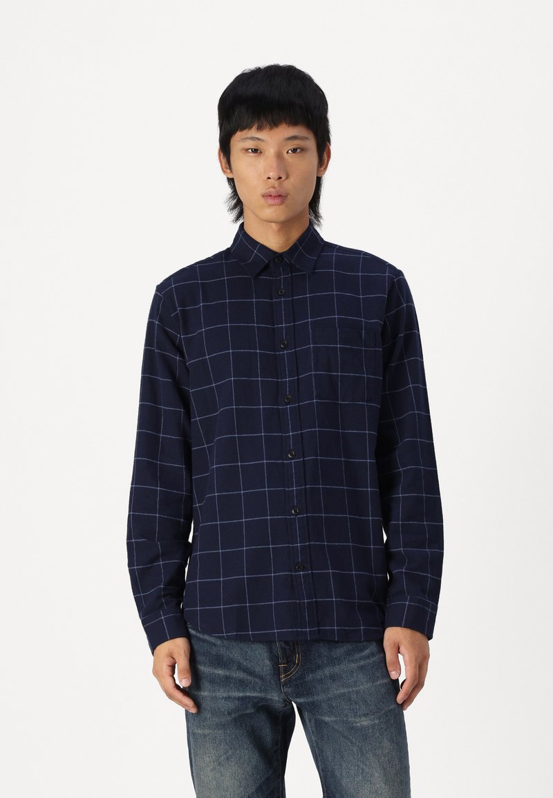 Camisa de cuadros marina hecha de algodón, con mangas largas, cuello abotonado y bolsillo frontal; presenta líneas de rejilla blancas y botones negros.