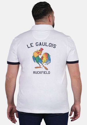 Homme portant un polo blanc avec bordure noire, un graphique coloré de coq, et les textes "LE GAULOIS" et "RUCKFIELD" au dos.