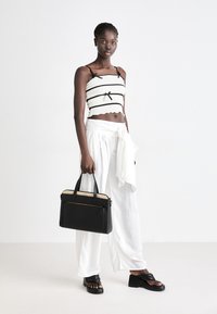 Borsa nera con accenti beige in rete, forma rettangolare e tasca anteriore con zip. Il modello indossa un top cropped bianco a righe e pantaloni bianchi larghi.