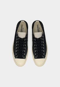 Ζευγάρι μαύρων και μπεζ χαμηλών αθλητικών παπουτσιών Converse με μαύρα κορδόνια, λευκή ραφή και λαστιχένιες μύτες σε ανοιχτό γκρι φόντο.