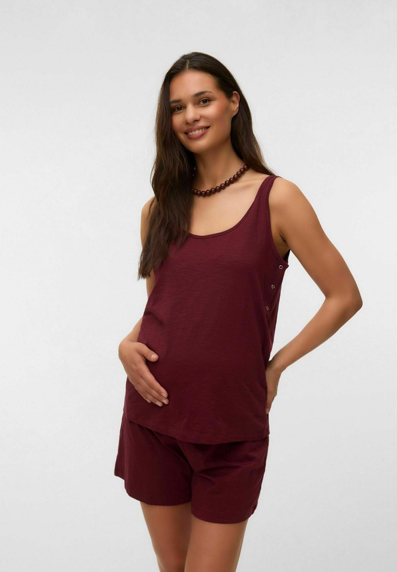 Femme enceinte portant un haut sans manches bordeaux et un short assorti, souriante, une main posée sur son ventre, devant un fond uni.