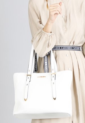 Bolso tote blanco con superficie texturizada, acentos de herrajes dorados y tirantes de logo en blanco y negro en contraste. Se muestra siendo sostenido en la mano.