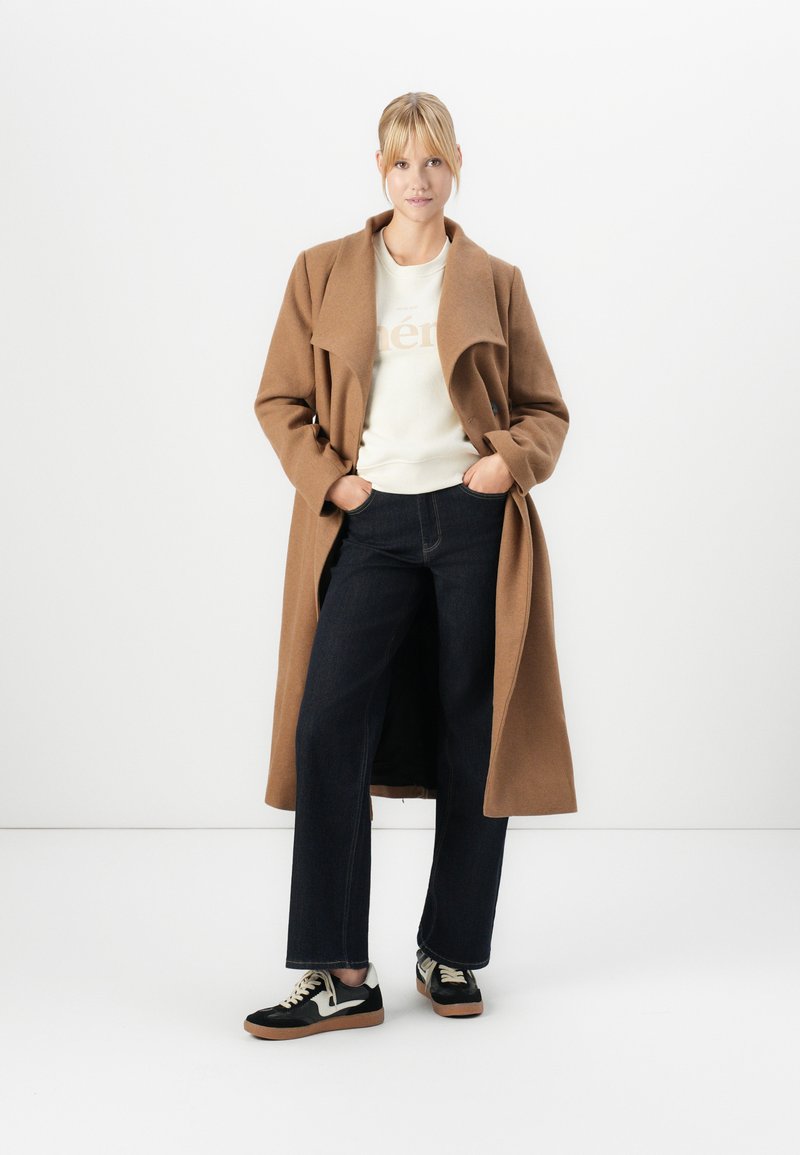 Lungo cappotto di lana color cammello con colletto ampio, indossato sopra un maglione leggero e jeans. Sneakers nere con dettagli crema e suola in gomma.