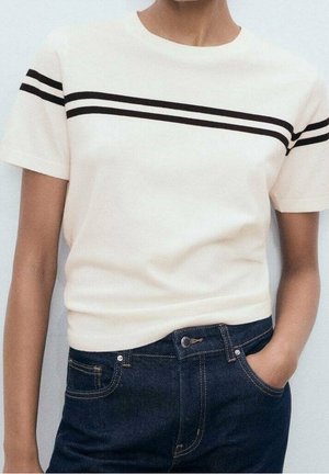 Person trägt ein weißes Kurzarmoberteil mit zwei schwarzen horizontalen Streifen über der Brust und den Schultern, kombiniert mit dunkelblauen Jeans.