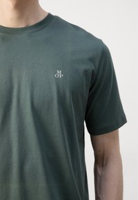 T-shirt en coton vert foncé avec un col rond, des manches courtes, et un petit logo blanc "M OP" sur la partie gauche de la poitrine. Texture douce.