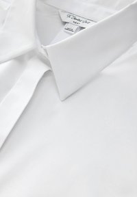 Bílá dlouhá košile s límcem, hladkou texturou, detail s knoflíkem na límci a viditelnou etiketou na výstřihu.