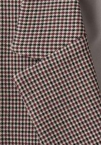 Geweven stof met een pied-de-poule patroon in bordeauxrood, zwart en crèmewit. Het materiaal heeft een textuurrijke uitstraling met een gladde afwerking.