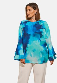 Blusa fluida a maniche lunghe in tonalità di blu e teal, con dettagli arricciati al collo e ai bordi delle maniche. Tessuto leggero.