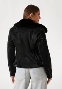 Schwarze Lederjacke mit einem Kunstfellkragen, gerippten Texturen an den Schultern, seitlichen Schnallen und silbernen Reißverschlüssen, kombiniert mit hellblauen Jeans.