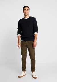 Maglione navy a coste, collo tondo bianco, pantaloni in corduroy verde oliva e sneakers bianche con suole marroni, in piedi su uno sfondo semplice.