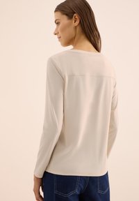 Beige long-sleeve top with a round neckline and a straight hem. Features a simple design with visible stitching along the back.