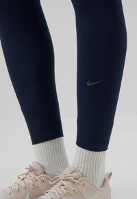Ben i marinblå leggings med Nike-logotyp, vita ribbade strumpor och ljusrosa Nike-sneakers mot en neutral bakgrund.