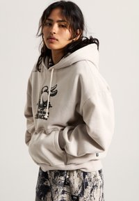 Beige hoodie with a graphic print, featuring a kangaroo pocket and drawstring hood. Patterns on black and white pants complement the outfit.