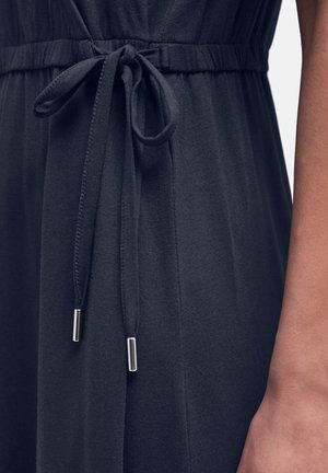 Cordon de serrage noué en un nœud sur une robe noire avec des embouts métalliques, présenté à côté d'un gros plan sur l'avant-bras et la main d'une personne.