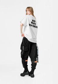 Camisa blanca de gran tamaño con "MOD WAVE MOVEMENT" impreso en negro. Pantalones cargo negros con múltiples bolsillos y acentos naranjas. Zuecos negros.