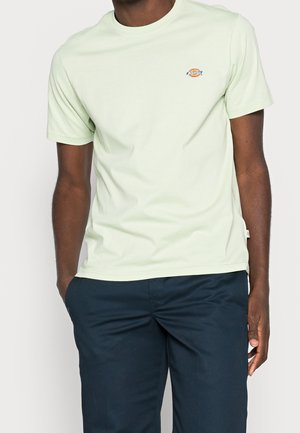 Hellgrünes Baumwoll-T-Shirt mit Rundhalsausschnitt und kurzen Ärmeln, mit einem kleinen, bunten Logo auf der linken Brust. Kombiniert mit marineblauen Hosen.