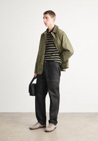 Khaki jacket, black striped shirt, dark denim pants, beige sneakers. Model holds a black handbag. Minimalist design, matte textures.