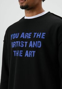 Schwarzes Sweatshirt mit Waffelstrickmuster, vorne mit großem, blauem Schriftzug "YOU ARE THE ARTIST AND THE ART". Weißer Rundhalsausschnitt.