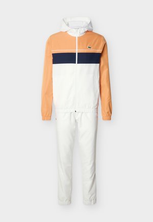 Herre hettejakke og joggedress med ferskenfargede ermer, hvit front og bukser, med en marineblå horisontal stripe og liten grønn krokodillelogo.