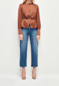 Blouse en satin bronze à manches longues, épaules froncées et taille cintrée, associée à un jean en denim bleu à jambe droite et des talons beiges.