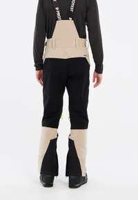 Beige en zwarte skibroeken met verstelbare bandjes, voorzien van ritsventilatie bij de enkels en een mix van texturen voor verbeterde functionaliteit.