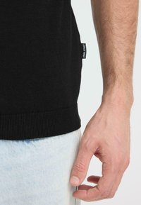 Pull en maille noire avec ourlet côtelé. Pantalon en denim bleu clair visible en bas. Petite étiquette de marque sur le côté du pull.