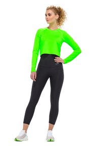 Neon zelený dlouhý rukávový cropped top v kombinaci s černými legínami s vysokým pasem. Má hladký materiál, přiléhavý střih a bílé sportovní boty se zelenými akcenty.