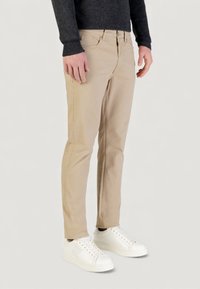 Pantalones chinos beige de corte ajustado, con cremallera y trabillas para cinturón, combinados con zapatillas blancas. Tela de algodón suave con detalles mínimos.