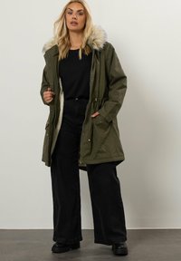 Olivgrön parkajacka med foder av päls i huvan, dragkedja och knäppning, elastisk midja och två framfickor. Bärs med svarta vida byxor.