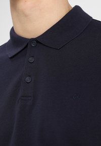 Polo shirt bleu marine foncé avec un col structuré, une patte de boutonnage à trois boutons et un logo CKJ brodé discrètement sur la poitrine. Tissu tricoté texturé.