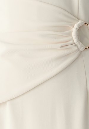 Witte stof met subtiele diagonale ribbeling, met gathering bij de taille, beveiligd door een gouden cirkelaccent.