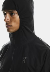 Giacca nera con collo alto e cappuccio, dotata di chiusura con zip e silhouette slim. Tessuto leggero con dettagli riflettenti.
