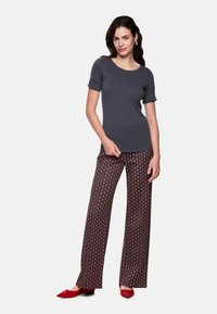 Graues Kurzarmoberteil kombiniert mit gemusterten, weit geschnittenen Hosen mit roten, orangefarbenen und cremefarbenen Rautenmuster. Rote flache Schuhe vervollständigen das Outfit.