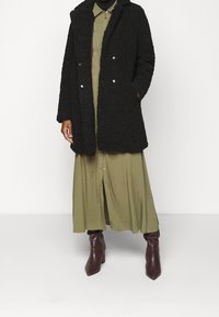 Cappotto in pelliccia ecologica nera con due bottoni, abbinato a un lungo vestito verde oliva e stivali al ginocchio marroni con tacco. Tessuto di una consistenza liscia.