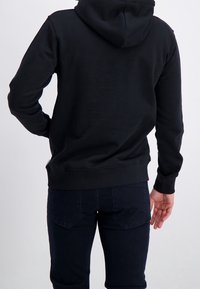 Personne portant un sweat à capuche noir et un jean foncé, debout avec une main sur la hanche et l'autre détendue le long du corps.