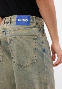 Világoskék farmernadrág kék címkével, rajta fehér "HUGO" felirattal, látható hátsó zsebekkel és enyhén kopott textúrával.