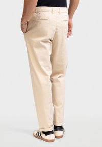 Man draagt beige opgerolde broek, zwarte top, zwarte sokken en witte sneakers met zwarte strepen, staat met één hand in zijn zak.