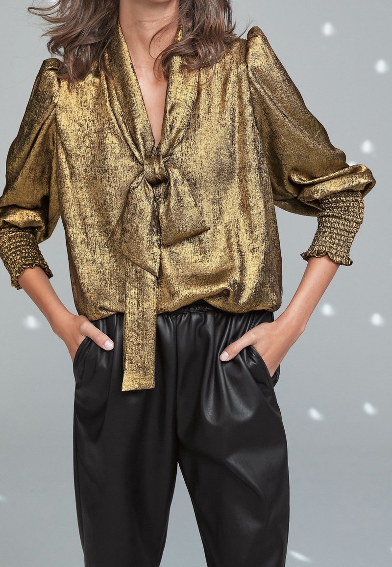Femme portant un chemisier doré métallique avec des manches bouffantes et un nœud au col, associé à un pantalon en cuir noir taille haute, mains dans les poches.